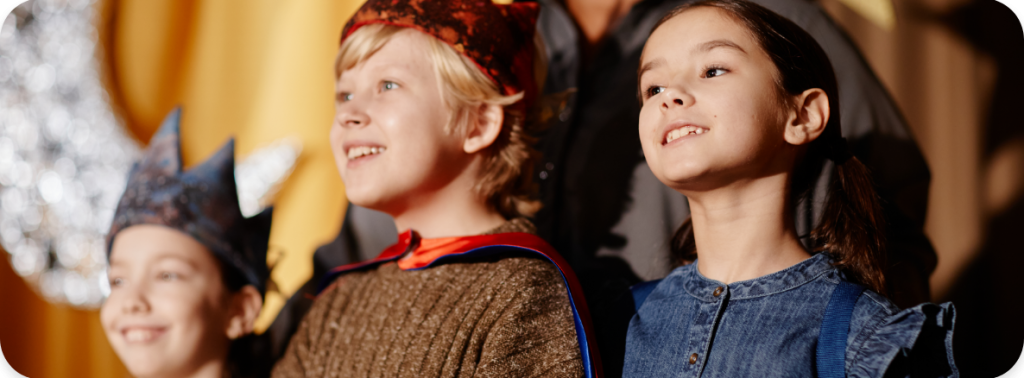 Trois enfants souriants costumés se tiennent sur scène pendant une pièce scolaire, dont deux portent des couronnes colorées. Un adulte ou enseignant se tient derrière eux, avec un rideau jaune et une lune et des étoiles en papier en arrière-plan.