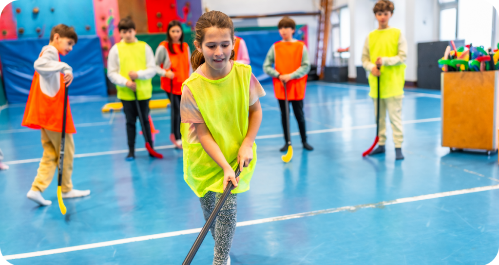 Un groupe d’enfants portant des dossards colorés joue au hockey intérieur dans un gymnase scolaire. Une fille au premier plan, portant un dossard jaune, manie un bâton de hockey rouge pendant que les autres observent ou attendent leur tour.