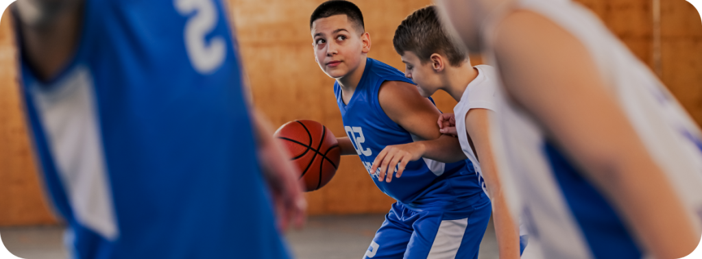 Jeunes joueurs de basketball en uniformes bleus et blancs s’affrontent dans un gymnase. Un joueur en bleu tient le ballon et cherche une occasion de passer ou de tirer, étroitement marqué par un adversaire en blanc.