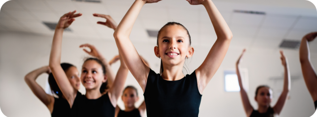 De jeunes filles dans un studio de danse, vêtues de tenues sportives noires, effectuent un grand écart assis tout en levant les bras en forme de cercle, dans une pose de ballet. La fille au centre sourit avec assurance pendant la routine de groupe.