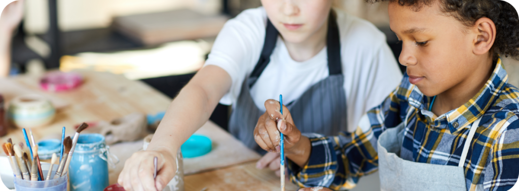 Deux enfants portant des tabliers travaillent sur un projet de peinture de poterie à une table couverte de pots de peinture et de pinceaux. Un enfant peint une petite tasse en céramique pendant que l’autre trempe un pinceau dans de la peinture bleue.